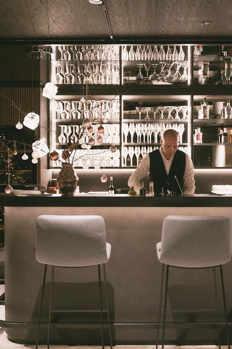 Erleben Sie einen Abend voller Genussmomente in Hinterstoder. Barkeeper bereitet Drinks im TRIFORÊT alpin.resort in Hinterstoder vor mit einem beeindruckendem Gläserregal im Hintergrund.