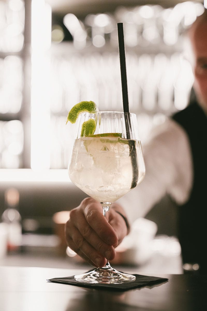 Cheers im Triforet: Ihr stilvoller Drink für besondere Abende. Nahaufnahme eines Cocktailglases mit Limette und Eiswürfeln an der Bar des TRIFORÊT alpin.resort in Hinterstoder.