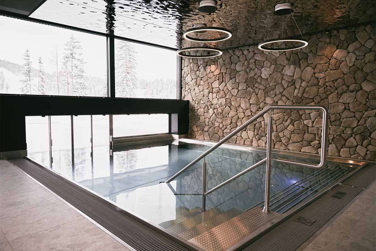 Entspannung mit Ausblick in unserem Indoorpool in Hinterstoder. Innenpool mit Natursteinwand, Deckenlichtern und Panoramafenster im TRIFORÊT alpin.resort in Hinterstoder.