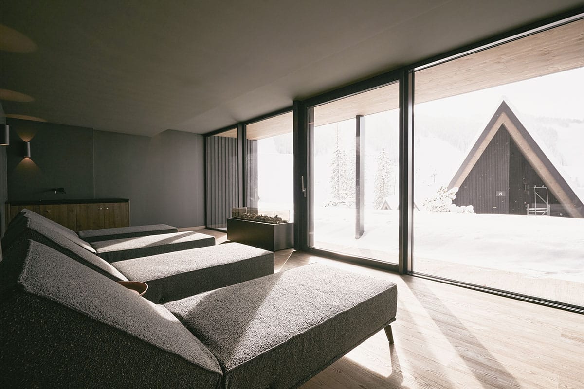 Ruhe mit Aussicht in unserem Entspannungsbereich in Hinterstoder. Moderner Ruheraum mit gepolsterten Liegen, Kamin und großen Fensterfront im Spa des TRIFORÊT alpin.resort.
