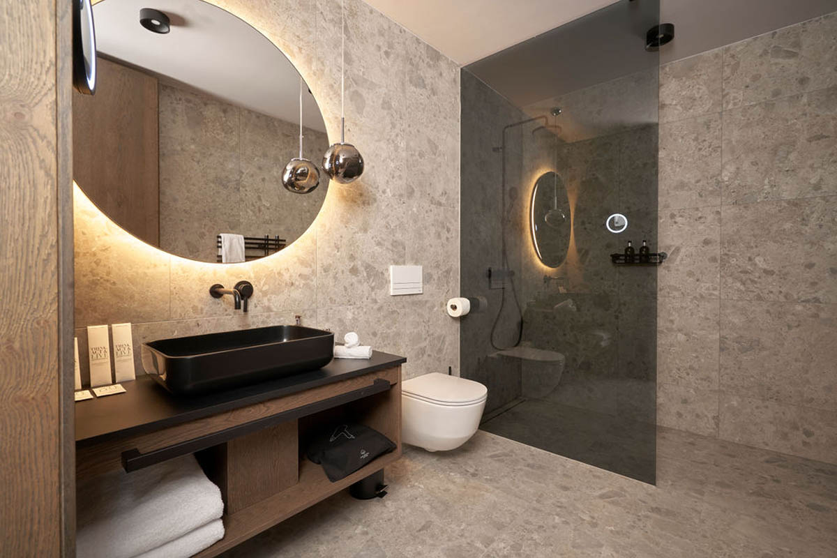 For your morning routine in style on vacation in the Alps. Bathroom with glass shower, round wall mirror, suspended WC and dark washbasin in the TRIFORÊT alpin.resort.