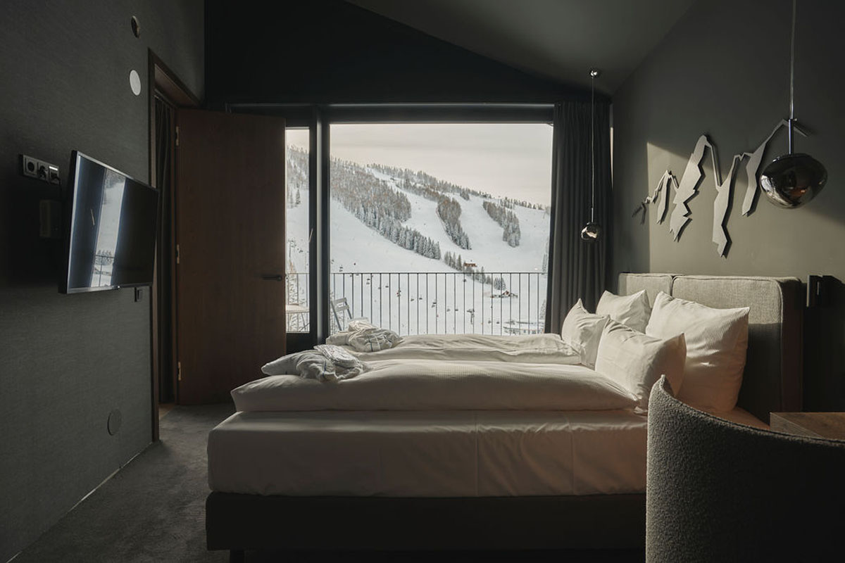 Fall asleep with a view on your vacation in the Alps. Double bed chalet of the TRIFORÊT alpin.resort with wall decoration, curtains and floor-to-ceiling window with mountain view.