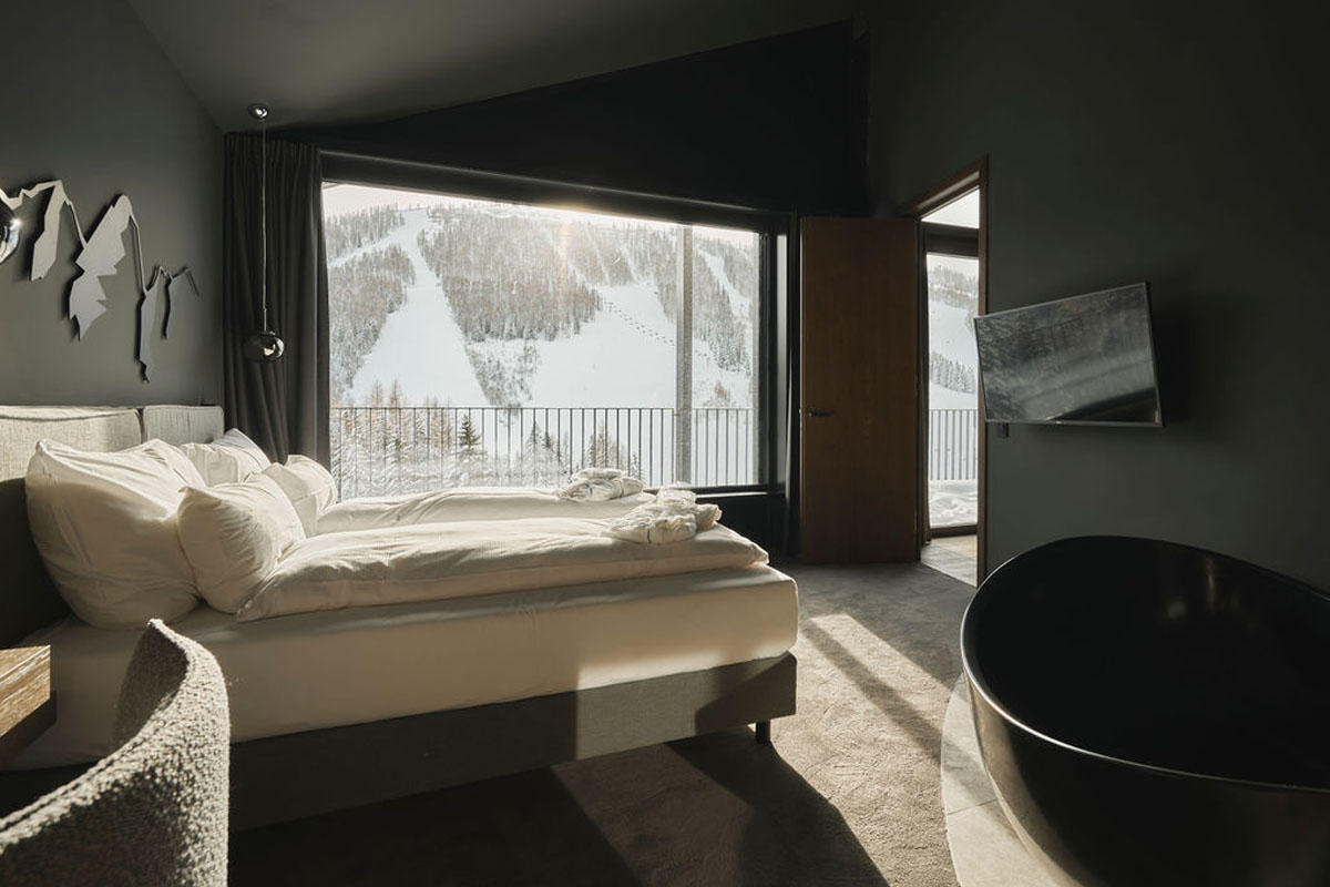 Wake up with a panoramic view - your bedroom on vacation in the Alps. Modern sleeping area with large window and view of snow-covered ski slopes in the TRIFORÊT alpin.resort in Hinterstoder.