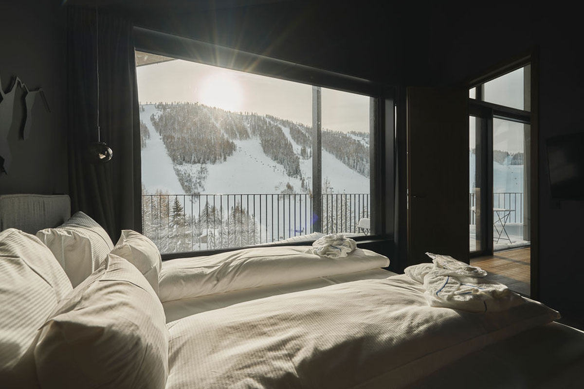 Morgensonne im Chalet – stilvolles Schlafzimmer mit traumhafter Aussicht. Modernes Schlafzimmer mit großen Fenstern und Blick auf verschneite Skipisten und Wald im TRIFORÊT alpin.resort in Hinterstoder.